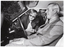 B&W photo of three men in or around a car. One man is seated in the driver's seat of the car. The car door is open, and the other two men are in the doorway, bending forward. All three men are smiling as they look at the steering wheel and dashboard of the car.