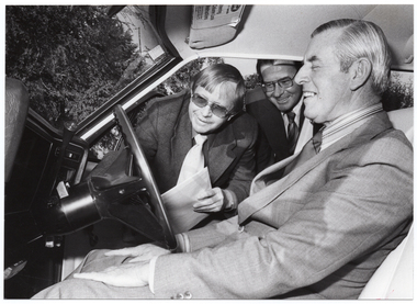 B&W photo of three men in or around a car. One man is seated in the driver's seat of the car. The car door is open, and the other two men are in the doorway, bending forward. All three men are smiling as they look at the steering wheel and dashboard of the car.