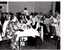 B&W photo of a large group of people sitting along several long rectangular tables in a large room. There are also a few people standing. In the background, the is a set of closed concertina doors. On one side of the concertina doors, there is a framed print on the wall, and one the other side, there is a large curtained window.