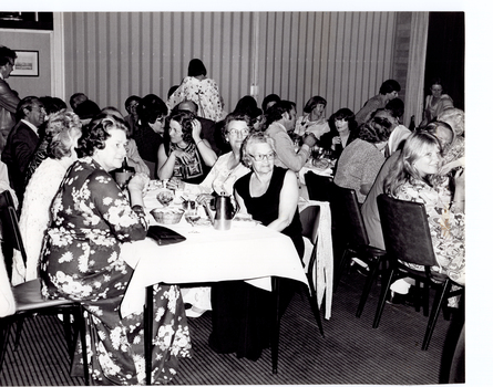 B&W photo of a large group of people sitting along several long rectangular tables in a large room. There are also a few people standing. In the background, the is a set of closed concertina doors. On one side of the concertina doors, there is a framed print on the wall, and one the other side, there is a large curtained window.