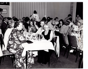 B&W photo of a large group of people sitting along several long rectangular tables in a large room. There are also a few people standing. In the background, the is a set of closed concertina doors. On one side of the concertina doors, there is a framed print on the wall, and one the other side, there is a large curtained window.