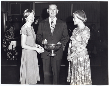 B&W photo of a man holding a large metal fruit bowl, from which raffle tickets are being drawn. Two women stand on either side of him. In the background, there are several people seated at tables. They are in a wood-panelled room with some large windows to one side.