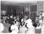 B&W photo of a large group of people gathered around a man, who is on one side of a large room. He is standing near the wall and a double doorway, facing the group of people in the room. There are various items hanging on the wall, including a notice board, a decorative wooden honour board, some coat hooks, and a clock.