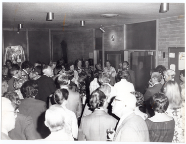 B&W photo of a large group of people gathered around a man, who is on one side of a large room. He is standing near the wall and a double doorway, facing the group of people in the room. There are various items hanging on the wall, including a notice board, a decorative wooden honour board, some coat hooks, and a clock.