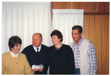 COL photo of four people, standing in a row, posing for a photo. Two women are holding a wad of cash and cheques (bound together with an elastic band), while two men look on. Behind them, there are some windows covered in white lace curtains and a wood-panelled wall.