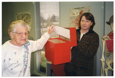 COL photo of an elderly woman and a younger woman. The elderly woman is holding an envelope above an open red cardboard box, while the younger woman is holding the box with its lid taken off. Beside the younger woman, there is a skeleton on a stand wearing a hat and a scarf (partially obscured). Someone's hand (partially obscured) is placed around the skeleton's shoulder/neck.