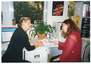 COL photo of two women, sitting at a desk, looking at a brochure which the woman on the left is holding.