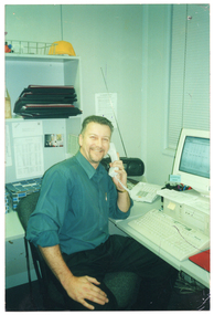 COL photo of a man smiling, while holding a telephone receiver up to his ear. He is sitting at a desk with a computer and keyboard on it.