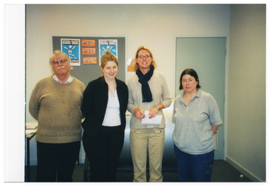 COL photo of four people, standing in a row, posing for a photo. One of them is holding a cheque and envelope. On the wall behind them are some National Healthy Bones Week posters.