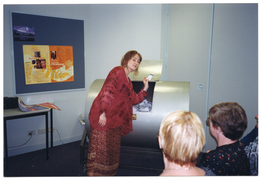 COL photo of a woman, standing. She is leaning towards a large open barrel affixed horizontally to a stand. She is holding a raffle ticket in one hand, and the barrel is full of raffle tickets. In the background, there is a display board on the wall with posters on it, and a small table against the wall, with a loose poster on top. In the foreground, there are a few people watching the woman.