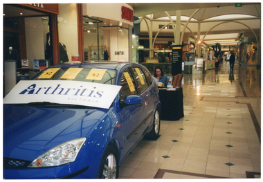 COL photo of a woman at a major shopping centre. She is in the background, sitting at a small table covered in a dark blue or black table cloth, behind a new blue car. There are various items on the table, including some printed leaflets. There is a wooden bench behind her. On the car's windscreen, there are three small (A4 size) yellow signs above one large white sign with 'Arthritis Victoria' in blue text. The letter 'A' in arthritis is a logo. Of the three A4 sized signs, two contain the text, "Tickets $2" - one placed on either side of the windscreen. The sign in the middle contains the text, "This Raffle is staffed by Volunteers". In the background, there are a variety of retail stores,  including a 'Santini' shoes, clothing and accessories store and a cafe.