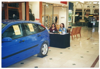 COL photo of two women at a major shopping centre. They are sitting at a small table covered in a dark blue or black table cloth, behind a new blue car. There are various items on the table, including printed leaflets and some plastic coin collection containers. Behind them, there is a large rubbish bin and a wooden bench. There is a small (A4 size) yellow sign on each of the car's two side windows. The first sign contains the text, "Only one week until Drawn!". The second sign contains the text, "Tickets $2". In the background, there are a variety of retail stores, including an unidentified a menswear store, a 'Santini' shoes, clothing and accessories store, a cafe, and a tie store called "Tie Rack".
