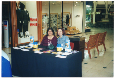 COL photo of two women at a major shopping centre. They are sitting at a small table covered in a dark blue or black table cloth, behind a new blue car. There are various items on the table, including raffle tickets, printed leaflets and factsheets, and some plastic coin collection containers. Behind them, there is a large rubbish bin and a wooden bench.