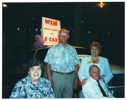 COL photo of four people, two sitting and two standing, in front of a new white car. The car has some large signs on it. One banner sign on the windscreen contains the text, "ARTHRITIS VICTORIA" (partially obscurred). An A-framed sandwich board sign on the roof of the car contains the text, "WIN YOUR CHOICE OF 2 CARS". The background is dark, with a few small lights.