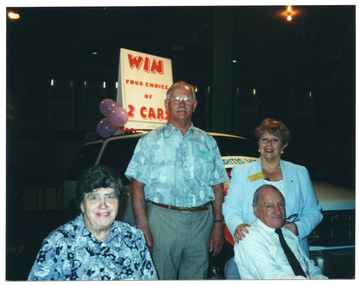 COL photo of four people, two sitting and two standing, in front of a new white car. The car has some large signs on it. One banner sign on the windscreen contains the text, "ARTHRITIS VICTORIA" (partially obscurred). An A-framed sandwich board sign on the roof of the car contains the text, "WIN YOUR CHOICE OF 2 CARS". The background is dark, with a few small lights.
