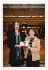 COL photo of two women standing in front of a department store and smiling. Together, they are holding an envelope which has a magenta and gold ribbon around it. In the top right corner of the envelope, the "MYER" logo/word is in large black text.