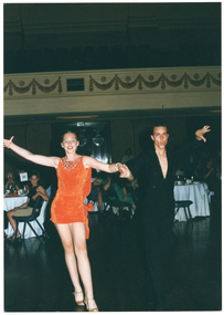 COL photo of two people, both facing the camera. They are clasping eath other's hand during a dance performance. Both are dressed in ballroom dancing costumes - the young man is wearing a black shirt and black trousers and the young woman is wearing an orange sleeveless mini dress. In the background, people are seated at large round tables covered in white tablecloths.