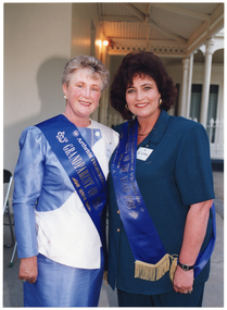 COL photo of two women posing for a photo on the veranda of a large white building. Each woman is wearing a royal blue sash (with silver lettering) across her body.