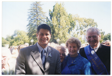 COL photo of four people posing for a photo outdoors. One of them is wearing a blue sash with gold text, "2001 Grandparent of the Year". In the background, there is a large crowd of people gathered behind them.