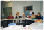 COL photo of a group of people seated at tables set in a circle formation. There are various items on the tables, such as folders and diaries, a lunchbox and several glasses, and a platter of apples. In the background, there is a telephone affixed to the wall, and a display board. There are several posters pinned to the display board, with headings such as "Active Living: get stronger with age", "National Healthy Bones Week", and "10 Steps for Living Well". Further along the wall, there is a set of closed double glass doors.