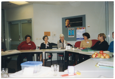 COL photo of a group of people seated at tables set in a circle formation. There are various items on the tables, such as folders and diaries, a lunchbox and several glasses, and a platter of apples. In the background, there is a telephone affixed to the wall, and a display board. There are several posters pinned to the display board, with headings such as "Active Living: get stronger with age", "National Healthy Bones Week", and "10 Steps for Living Well". Further along the wall, there is a set of closed double glass doors.