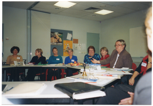 COL photo of a group of people seated at tables set in a circle formation. There are various items on the tables, such as a small brass bell, folders, diaries and note books, a lunchbox and glasses, and a platter of apples. In the background, there is a telephone affixed to the wall next to a display board. There are posters pinned to the display board, with headings such as "Active Living Age is no barrier to good health" and "The Pyramid of Good Health".  In the centre-left of the frame, there is a set of large bi-fold doors folded back in the open position. To the right of the frame, there is a large whiteboard affixed to a perpendicular wall.