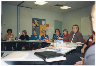 COL photo of a group of people seated at tables set in a circle formation. There are various items on the tables, such as a small brass bell, folders, diaries and note books, a lunchbox and glasses, and a platter of apples. In the background, there is a telephone affixed to the wall next to a display board. There are posters pinned to the display board, with headings such as "Active Living Age is no barrier to good health" and "The Pyramid of Good Health".  In the centre-left of the frame, there is a set of large bi-fold doors folded back in the open position. To the right of the frame, there is a large whiteboard affixed to a perpendicular wall.