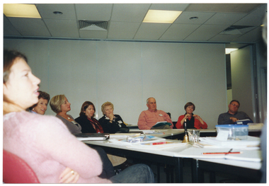COL photo of a group of people seated at tables set in a circle formation. There are various items on the tables, such as a small brass bell, folders, diaries and note books, a pencil, a glass, and a lunchbox. In the background, there is a set of large bi-fold doors held in the closed position, with the last panel opening out into the room behind.
