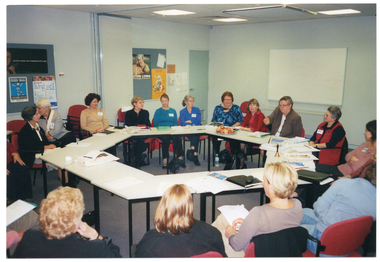 COL photo of a group of people seated at tables set in a circle formation. There are various items on the tables, such as a small brass bell, folders, diaries, note books, leaflets, pens and pencils, a glass, a lunchbox, and a platter of apples. In the background are two display boards affixed to the wall, with posters pinned on the boards. The posters on one board contain headings such as "Active Living: get stronger with age", "National Healthy Bones Week", and "10 Steps for Living Well". The posters on the other board contain headings such as "Active Living Age is no barrier to good health", and "The Pyramid of Good Health". In the centre-left of the frame, there is a set of large bi-fold doors folded back in the open position. Further along the wall, there is a closed door. To the right of the frame, there is a large whiteboard affixed to a perpendicular wall.