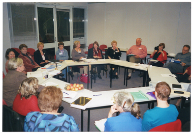 COL photo of a group of people seated at tables set in a circle formation. There are various items on the tables, such as a small brass bell, folders, diaries, note books, leaflets, pens and pencils, a lunchbox, and a platter of apples. In the background, there is a set of double glass doors with a large window on either side. The windows have venetian blinds attached.
