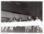 B&W photo of five people sitting at a long table on a stage. Two microphones on stands have been placed on the table, along with two jugs of water and some glasses. In the background, an empty section of balcony-level seating is evident. The photo has been taken from a low angle.