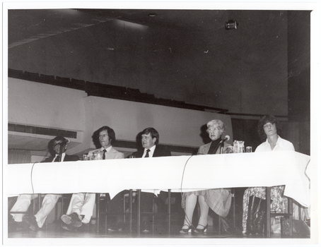 B&W photo of five people sitting at a long table on a stage. Two microphones on stands have been placed on the table, along with two jugs of water and some glasses. In the background, an empty section of balcony-level seating is evident. The photo has been taken from a low angle.