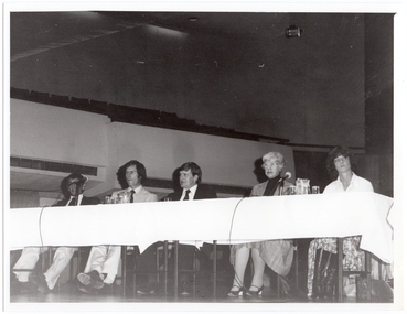 B&W photo of five people sitting at a long table on a stage. Two microphones on stands have been placed on the table, along with two jugs of water and some glasses. In the background, an empty section of balcony-level seating is evident. The photo has been taken from a low angle.