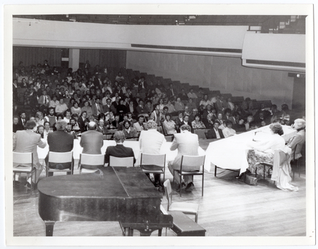 B&W photo of eight people sitting at two long tables on a stage. There are six people on one table and two people evident sitting on the second table (which is partially obscured). Two microphones on stands are evident on the second table. There is a large piano in the foreground, behind one of the tables. In the background, a large audience, who are seated on rows of seats, fills the auditorium. At the top of the frame, several sections of empty balcony seating are evident. The photo has been taken from a height, at a slight angle.