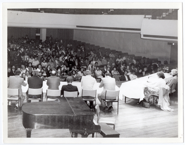 B&W photo of eight people sitting at two long tables on a stage. There are six people on one table and two people evident sitting on the second table (which is partially obscured). Two microphones on stands are evident on the second table. There is a large piano in the foreground, behind one of the tables. In the background, a large audience, who are seated on rows of seats, fills the auditorium. At the top of the frame, several sections of empty balcony seating are evident. The photo has been taken from a height, at a slight angle.