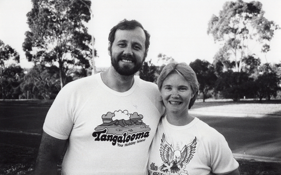 B&W medium close up photo of a man and a woman standing outdoors. He has one arm around her shoulders, and they are both smiling. He is wearing a t-shirt with a picture and text that says, "Tangalooma Your Holiday Island". She is wearing a t-shirt with some text (obscured) and a graphic drawing of an eagle. Behind them, there is an empty street with several trees in a park across the road.