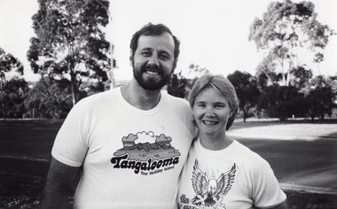 B&W medium close up photo of a man and a woman standing outdoors. He has one arm around her shoulders, and they are both smiling. He is wearing a t-shirt with a picture and text that says, "Tangalooma Your Holiday Island". She is wearing a t-shirt with some text (obscured) and a graphic drawing of an eagle. Behind them, there is an empty street with several trees in a park across the road.