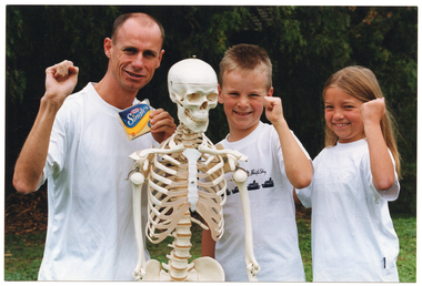 COL photo of a man and two children standing next to a plastic skeleton on a stand. They are each holding one fist up and smiling. The man is also holding up a packet of Kraft Singles cheese slices in his other hand. They are standing outdoors, with some leafy bushes in the background.