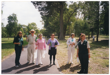 COL photo of a small group of people of varying ages posing for a photo along a flat concrete path at a park. Several people in the group are holding up a branded green plastic water bottle. One person has a dog on a leash (partially obcured).