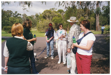COL photo of a small group of people of varying ages standing on a flat concrete path at a park, with two park rangers. Several people in the group are holding up a branded green plastic water bottle. One person has a dog on a leash.