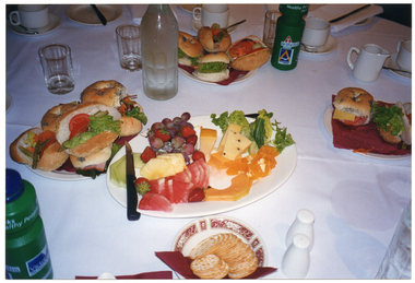 COL photo of various items of food. set up on a table covered with a white tablecloth. The food consists of salad rolls, a fruit and cheese platter, and a small plate of crackers. Also on the table is a glass bottle of water, two 'Healthy Parks Healthy People' branded green plastic water bottles, some glasses and various pieces of white crockery (cups and saucers, a small white milk jug, and a set of salt and pepper shakers).