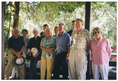 COL photo of a group of people of varying ages posing for a group photo under a shelter, behind some wooden benches. In the background, there are several trees covered in green foliage.