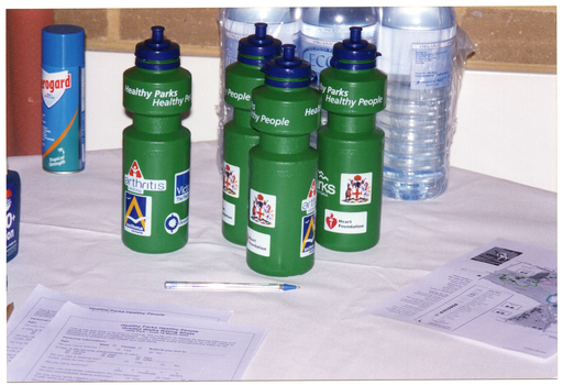 COL photo of various items on a table covered with a white tablecloth. There are several 'Healthy Parks Healthy People' branded green plastic water bottles, some clear plastic water bottles, a can of Aeroguard mosquito spray, a bottle of sunscreen (partially obscured), a pen, some survey forms, and a printed flyer showing a map.