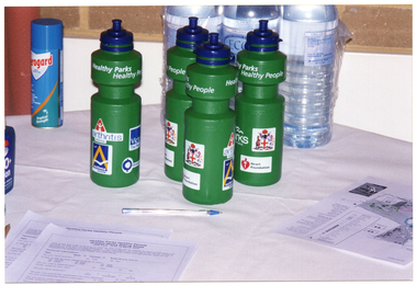 COL photo of various items on a table covered with a white tablecloth. There are several 'Healthy Parks Healthy People' branded green plastic water bottles, some clear plastic water bottles, a can of Aeroguard mosquito spray, a bottle of sunscreen (partially obscured), a pen, some survey forms, and a printed flyer showing a map.