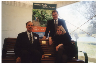 COL photo of three people posing for a photo inside a marquee. One man and a woman are sitting on a wooden bench, while another man is standing behind the bench. A small engraved brass plaque has been affixed to the bench. Behind them is a vertical free-standing banner titled "Healthy Parks Healthy People".