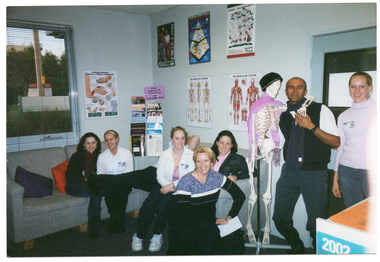 COL photo of a group of people posing for a photo with a plastic skeleton on a stand.  Some people are seated on two grey sofas, while two others are standing near the skeleton. On the walls are several posters, with headings such as 'Understanding carpel tunnel syndrome', 'The skeletal system', and 'The muscular system'. In the corner, there is a display stand with magazines and information sheets on it. Attached to the display stand is a sign which says "These journals and newsletters are NOT FOR LOAN".