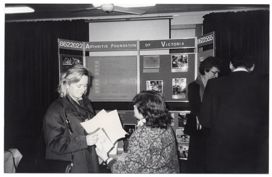 B&W photo of four people standing in front of a multipanel display board. Displayed across the top of the centre board is the text, "ARTHRITIS FOUNDATION OF VICTORIA", with the phone number "8622022" across the top of the two side panels.
