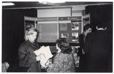 B&W photo of four people standing in front of a multipanel display board. Displayed across the top of the centre board is the text, "ARTHRITIS FOUNDATION OF VICTORIA", with the phone number "8622022" across the top of the two side panels.