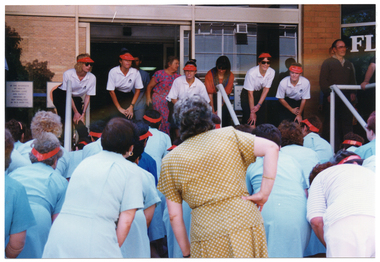 COL photo of a group of people demonstrating some exercises to a group of nurses. The people demonstrating the exercises are on an open veranda outside a tan brick building, while the nurses are in the forecourt or ground below. There are a few people standing on the veranda, to the right of the frame.
