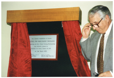 COL photo of a man standing beside an engraved plaque.  The plaque is on a display board covered with black fabric. Above the board is a wooden pelmet, beneath which are two short red curtains that have been drawn open. Engraved on the plaque is the text, "This Arthritis Foundation of Victoria CENTRE FOR RHEUMATIC DISEASES Incorportating the H & L Hecht Visitiing Professorship was officially opened by Professor Sir Gustav Nossal, AC, CBE on 2nd March 1995". Below this is some additional text (partially obscured) on the bottom left: "...ject of", "Foundation of Victoria Inc.", "of Melbourne", and "...lbourne Hospital". On the bottom right of the plaque is the text: "President: Dr. Les Koadlow", "Medical Dean: Professor Graeme Ryan, AC", "President: Hon. Margaret Guilfoyle, DBE".